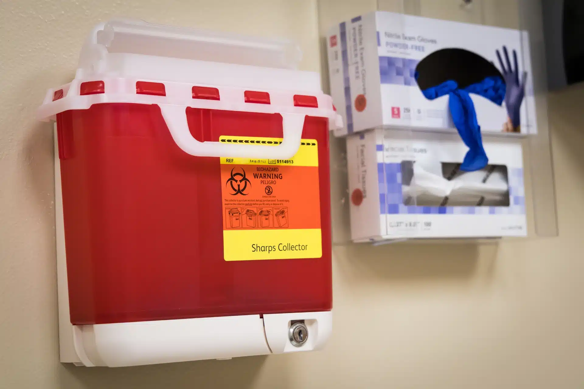 A red wall-mounted sharps disposal container with a biohazard warning label is shown beside boxes of medical gloves and tissues in a clinical setting 