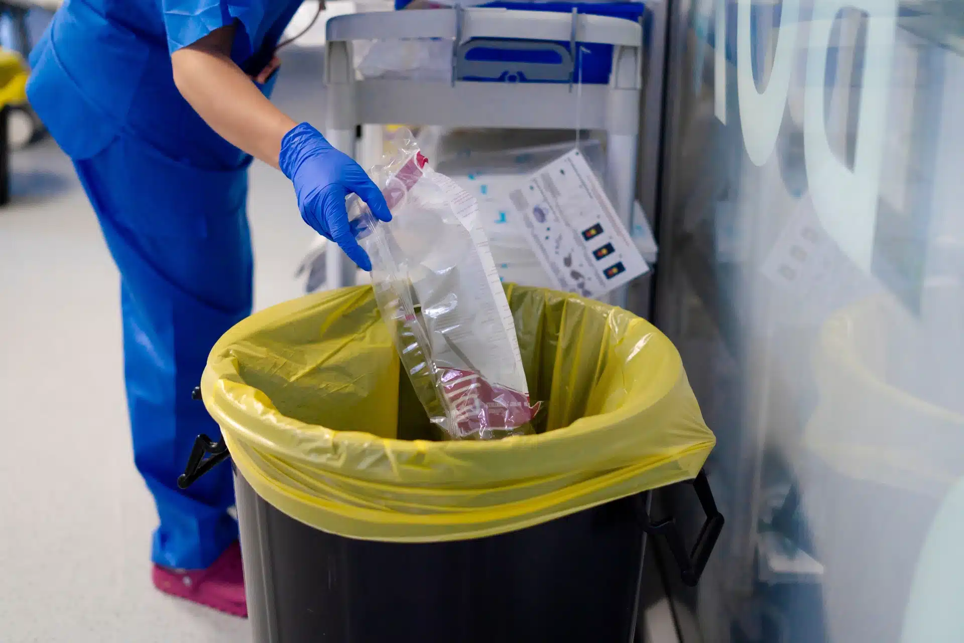 Healthcare worker in blue scrubs and gloves disposing of medical waste into a yellow-lined bin in a clinical setting