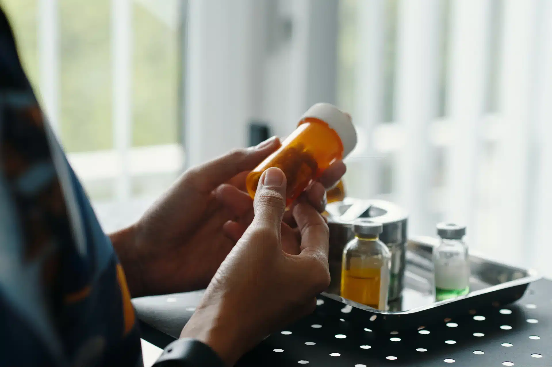 Close-up of a person holding an amber pill bottle, with small medical vials sitting on a metal tray in the background. 